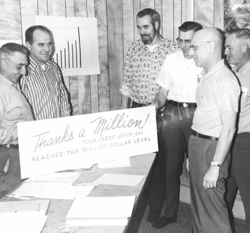 Black-and-white photo of five men standing around a desk in an office, smiling and looking at a large sign that reads “Thanks a Million! Your credit union has reached the million dollar level.” Papers are spread across the desk, and a simple bar chart is displayed on the wood-paneled wall behind them.
